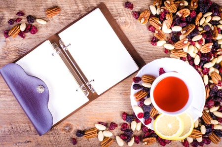 Dried fruits - pecan, cranberry, raisin, almond with white cup of tea and  open notebook on wooden background.の写真素材