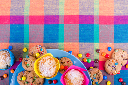 Fresh homemade muffins and cookies with color chocolate coated candies on a colorful checkered kitchen towel. Free space for your textの写真素材