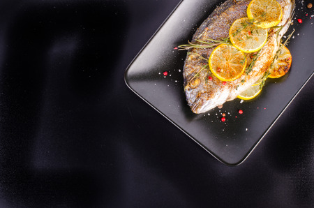 Roasted gilthead fishes with lemon, herbs, salt on black background. Healthy food concept. Food frame. Free space for your text.の写真素材