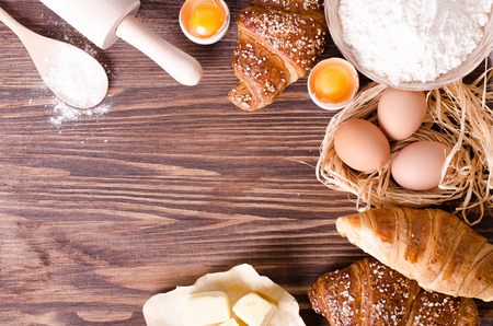 Ingredients for baking croissants - paper, flour, wooden spoon, rolling pin, eggs, egg yolks, butter served on a rustic wooden tray table.の写真素材