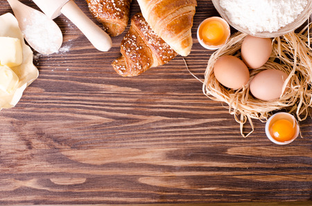 Ingredients for baking croissants - paper, flour, wooden spoon, rolling pin, eggs, egg yolks, butter served on a rustic wooden tray table.の写真素材