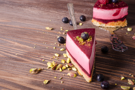 Piece of delicious raspberry cake with fresh raspberries, blueberry, currants and pistachios on shovel, wooden background. Free space for your text.の写真素材