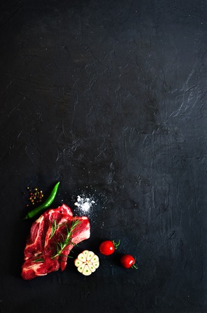 Raw fresh meat steak with cherry tomatoes, hot pepper, garlic, oil and herbs on dark stone, concrete background. Free space for your textの写真素材