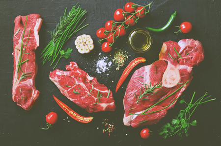Raw meat steaks with cherry tomatoes, hot pepper, garlic, oil and herbs on dark stone, concrete background. Free space for your textの写真素材