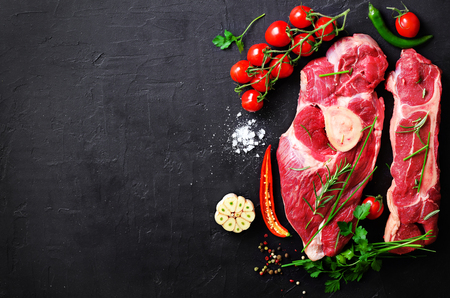 Raw meat steaks with cherry tomatoes, hot pepper, garlic, oil and herbs on dark stone, concrete background. Free space for your textの写真素材