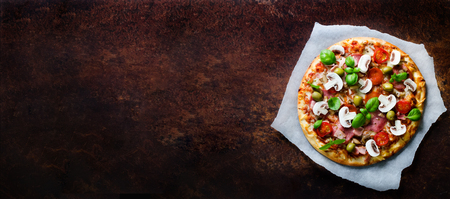 Fresh italian pizza with mushrooms, ham, tomatoes, cheese, olive, basil on backing paper, dark rustic background. Copy space. Homemade with love. Fast delivery. Recipe and menu. Bannerの写真素材