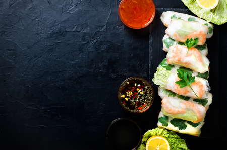 Fresh Vietnamese, Asian, Chinese food frame on black concrete background. Spring rolls rice paper, lettuce, salad, vermicelli, noodles, shrimps, fish sauce, sweet chili, soy, lemon. Top view.の写真素材