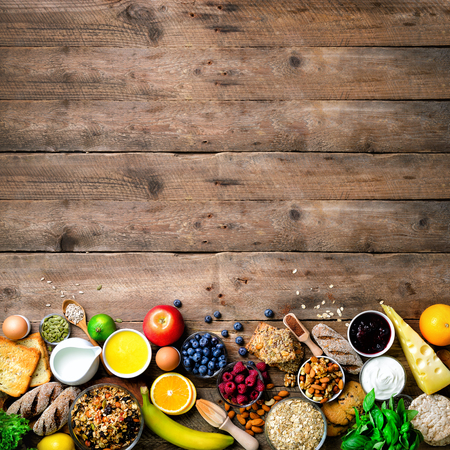 Healthy breakfast ingredients, food frame. Granola, egg, nuts, fruits, berries, toast, milk, yogurt, orange juice, cheese, banana, apple on wooden rustic background, top view, copy spaceの写真素材