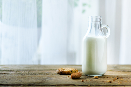 Chocolate chip cookies, bottle and glass of milk on wooden table near window, white background. Sunny morning, copy space.の写真素材