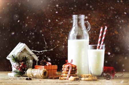 Christmas dark background with snow and bokeh, copy space. Bottle, glass with milk for Santa, cookies, red rope, anise stars, cinnamon sticks, cones, decor on wooden vintage tableの写真素材