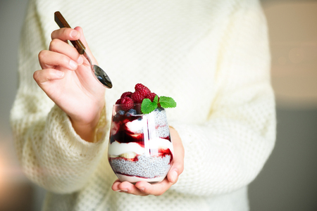 Girl in white sweater holds glass with chia milk dessert, raspberries, blueberries, mint. Healthy breakfast concept. Copy space, close up. Sunny morningの写真素材