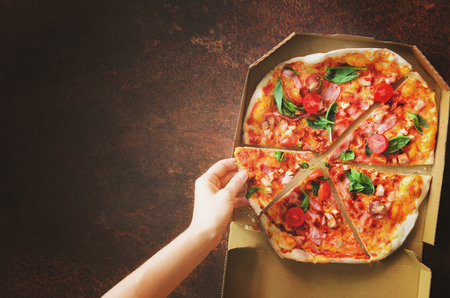 Female hand taking slice of fresh pizza from delivery box. Top view, dark background. Junk food. Sunny morning with sunlight. Toned pastel colorの写真素材