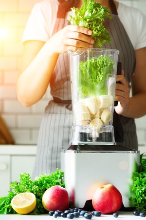 Woman blending lettuce leaves, spinach, aplles, berries, bananas. Homemade healthy green smoothie. White kitchen design. Sunny morning with sunlight. Toned effectの写真素材