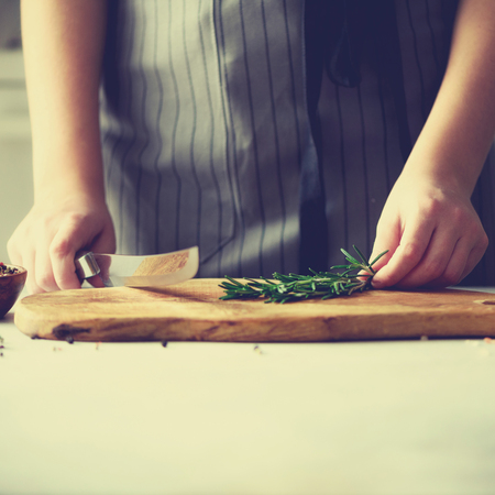 Woman hands cutting fresh green rosemary on wood chopping board in white kitchen, interior.の写真素材