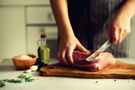 Woman hands cutting beef meat on wooden chopping board, rosemary, oil, salt, pepper. Girl cooking pork meat on white kitchen background.の写真素材