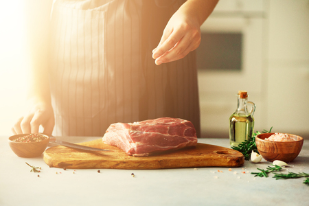 Woman hands cutting beef meat on wooden chopping board, rosemary, oil, salt, pepper. Girl cooking pork meat on white kitchen background.の写真素材