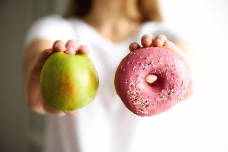 Young woman in white T-shirt choosing between green apple or junk food, donut. Healthy clean detox eating concept. Vegetarian, vegan, raw concept. Copy space.の写真素材