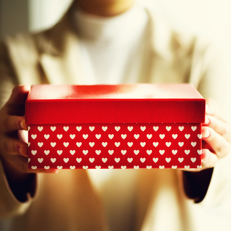 Female hands holding red gift box with white hearts, copy space. Christmas, hew year, birthday, valentines day concept. Square crop.の写真素材