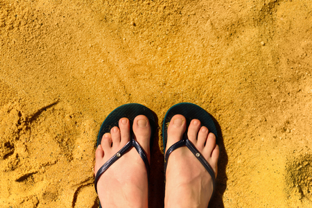 Woman legs in slippers on yellow sand background. Blue flip flops on beach. Copy space, top view....