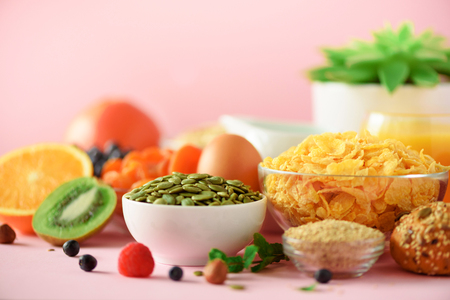 Corn flakes with milk, pumpkin seeds, fresh berries, yogurt, boiled egg, nuts, fruits, orange, banana, peach for breakfast on pink background. Copy space. Healthy food conceptの写真素材