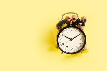 Hand holding black alarm clock through hole in yellow paper