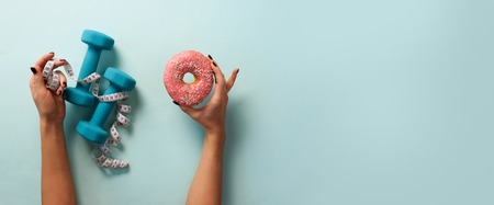 Female hand holding sweet donut, measuring tape, dumbbells over blueの写真素材