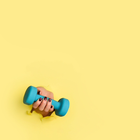 Woman hand holding blue dumbbell on yellow background. Fitness, sport, healthy lifestyle, diet concept. Banner with copy space. Square crop.の写真素材