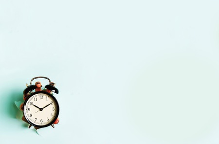 Hand holding black alarm clock through hole in blue paper background. Wake up alert concept. Morning routine. Banner with copy space.の写真素材