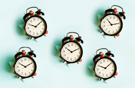 Chaotic pattern of hand holding black alarm clock through hole in blue paper background. Wake up...