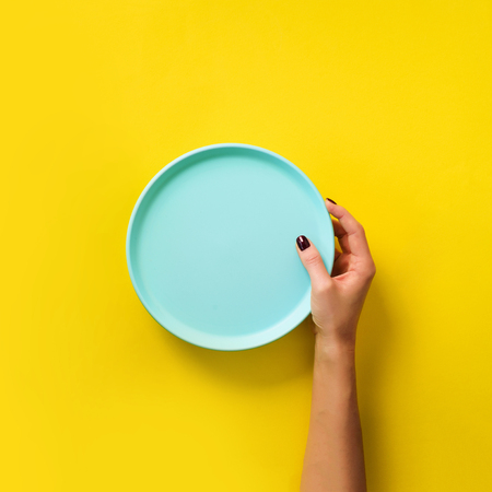 Female hand holding empty blue plate on yellow  with copy space. Healthy eating, dieting concept. Square crop.の写真素材