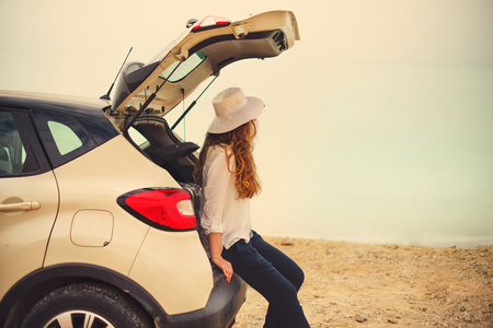 Happy stylish young woman traveler on beach road sitting on white crossover car, holding hat in hand.の写真素材