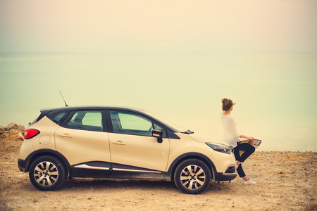 Happy stylish young woman traveler on beach road sitting on white crossover car, holding hat in hand.の写真素材