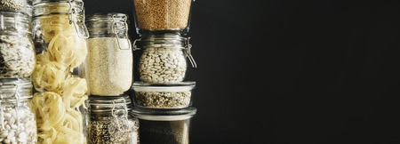 Banner with assortment of grain products and pasta in glass storage containers on wooden table....