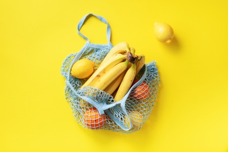 Mesh shopping bag with organic banana, orange, lemons on yellow paper background. Flat lay, top...