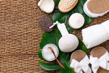 Skincare, beauty and spa concept with bath cosmetic accessories on rattan background. Flat lay. Top view of white towel, bamboo slippers, cream, wooden brush, coconut oil, monstera leaves.の写真素材