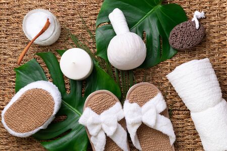 Spa, wellness, skin care, beauty and relax concept. Spa tools: white towel, bamboo slippers, herbal ball, cream, wooden brush, coconut oil, monstera on rattan backgroundの写真素材