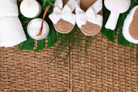 Spa tools: white towel, bamboo slippers, herbal ball, cream, wooden brush, coconut oil, monstera on rattan background. Cosmetic products for body treatment. Beauty, relax and massage concept.の写真素材