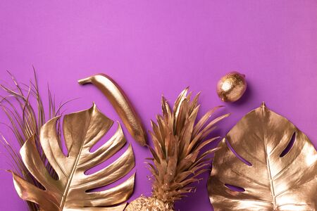 Golden exotic fruits, tropical palm, monstera leaves on violet background. Top view. Flat lay. Food concept. Creative layout of gold pineapple, banana, lemon with copy spaceの写真素材