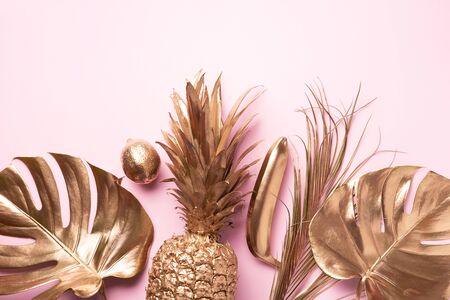Golden exotic fruits, tropical palm, monstera leaves on pink background. Top view. Flat lay. Food concept. Creative layout of gold pineapple, banana, lemon with copy spaceの写真素材