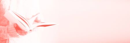 Young girl in white cardigan reading opened book. Copy space. Lifestyle and school concept in trendy coral color. Banner.の写真素材
