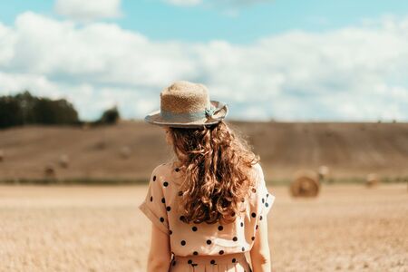 Woman in beige polka dot dress with curly hair, straw hat standing on harvested field with straw...