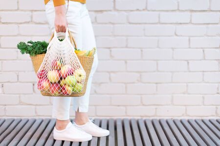 Girl holding mesh shopping bag full of apples and straw bag with organic vegetables, brick background. Zero waste, plastic free concept. Sustainable lifestyle. Copy space.の写真素材