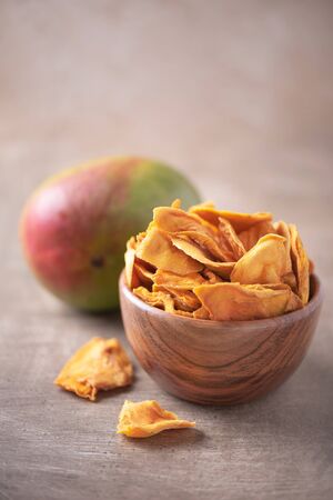 Dried mango in wooden bowl and fresh mango fruit on wood textured background. Copy space. Superfood, vegan, vegetarian food concept. Macro of orange dried mango slices, selective focus. Healthy snackの写真素材