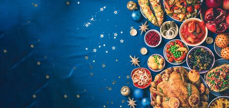 Roasted Christmas turkey with orange slices, cranberries, garlic, festive decoration, candles, tangerine, pomegranate, golden cultery, star glitter sparkles on blue background. Top view, copy space. Festive family Christmas dinnerの写真素材