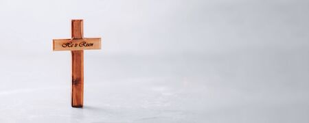 Wooden cross with text He is risen on grey background. Reminder of Jesus sacrifice and Christ resurrection. Easter passover. Eucharist concept. Christianity symbol and faith.の写真素材