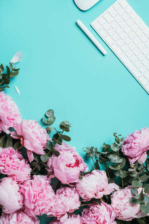 Home office workspace with white keyboard, notebook and peony, eucalyptus flower, copy space on blue background. Feminine workspace. Flat lay. Blogger, business concept. Mock up. Birthday, Woman day.の写真素材