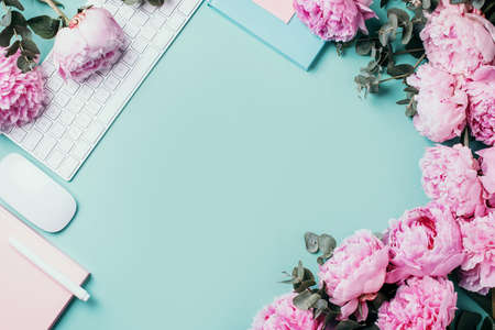 Home office workspace with white keyboard, notebook and peony, eucalyptus flower, copy space on blue background. Feminine workspace. Flat lay. Blogger, business concept. Mock up. Birthday, Woman day.の写真素材