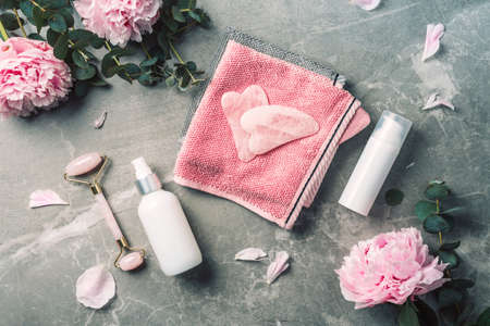 Facial roller, gua sha massager, oil bottle, towel, pink peonies on marble background. Copy space. Flat lay. Top view. Routine facial skin care, body treatment, spa concept.の写真素材