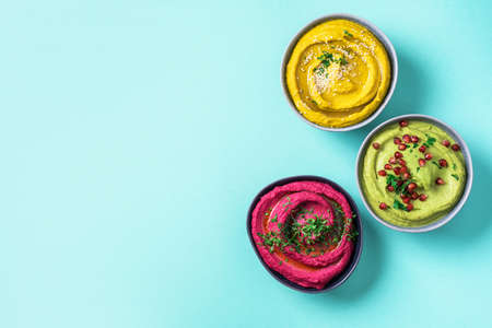 Colorful hummus bowls - green, yellow and beetroot hummus on dark background with lemon, olive oil, sesame seeds, pita, raw chickpeas, avocado, pomegranate. Middle eastern, jewish, arabic cuisine.の写真素材