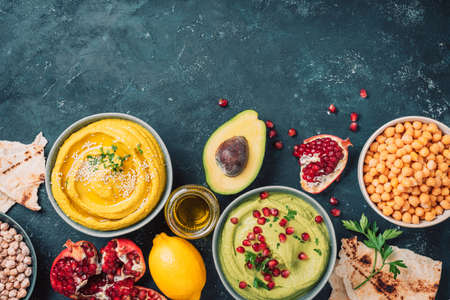 Ingredients for yellow hummus and green hummus - chickpea, tahini, olive oil, sesame seeds, pita, avocado, pomegranate on dark background. Middle eastern, jewish, arabic cuisine. Top view. Copy spaceの写真素材
